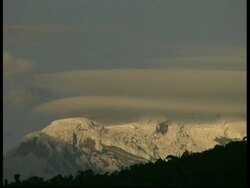 WA snow covered Mount Chimborazo, sunlit snow covered mountain peak with low bands of grey cloud in blue sky, silhouetted tree lined foreground, Ecuador Stock Footage