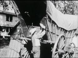 MOT: EARLY AMERICANA: WESTWARD EXPANSION REENACTMENT: Male, father loading large wooden barrel into Conestoga covered wagon, attaching back panel. Instructional Video