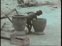 Two naked, starving children stand near large, clay pots. News Clip