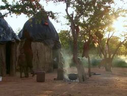 MS Child sitting on ground in front of lares with smoke around and woman moving hut side / Imba Village, Namibia Stock Footage
