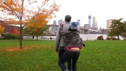 Young woman jumps on boyfriend's back and spins in autumn park; Brooklyn Bridge and New York skyline in background Stock Footage