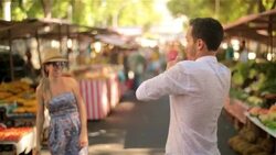 Girl blows boyfriend a kiss which he catches and mimics a beating heart in sunny marketplace Stock Footage