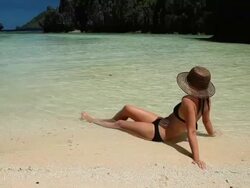 WS, beautiful caucasian woman in bikini and sun hat sitting on beach at water's edge relaxing at tropical secluded lagoon / Matinloc Island, Bacuit Archipelago, El Nido, Palawan, Philippines Stock Footage