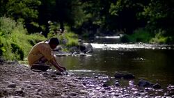 Female Native American Indian squatting by riverbank washing bowls in water with rocks in foreground Stock Footage