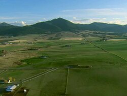 Aerial wide side-looking  POV of green farm fields and ranches near Bozeman, MT with mountains behind Stock Footage