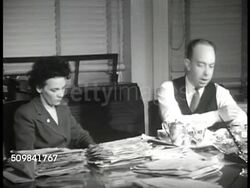 1946: WASHINGTON, D.C: NIGHT: People walking FG w/ clock at 9:00 pm BG. EXT Building w/ upper floor lights on. Wilson Wyatt working at desk w/ secretary, looking over paperwork, eating sandwich. Newspaper headline: 'Nearly All Building...Forbidden... Instructional Video