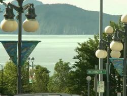"View from 4th Avenue of Resurrection Bay water, forested mountains behind. Lamposts with round lamps and ""SEWARD"" banners visible. Seward, Kenai Peninsula, Alaska." Stock Footage