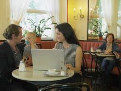 WS young woman sitting at table in traditional coffee house, writing on laptop computer; mature woman in background reading newspaper; young man carrying baby enters frame right, kisses young woman and takes seat at table; they talk Stock Footage