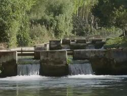 man in wading trousers carrying fish net walking across weir in fish farm, looking into water checking for fish, RED R3D 4K,4KMSTR Stock Footage