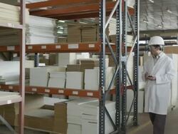 TS man in lab coat and hard hat entering frame right and  walking along multi-story warehouse in furniture production, taking notes on handheld electronic device; as clip advances, man comes progressively closer to camera (starts as WS, ends as CU) Stock Footage