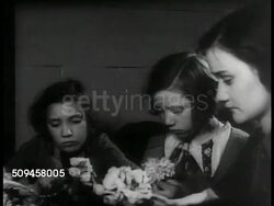 1937: CHILD URBAN WORK: Girls sitting at table w/ adult woman wrapping florist wire around silk bouquets. Girls working at table w/ netting & small black dots, Garment District 'Sweat Shop' work. Instructional Video