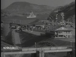 1940: PANAMA CANAL ZONE: HA WS Panama city houses w/ water BG. WS US Warship moving into canal. Destroyer moving slowly in lock of canal. Behind two soldiers standing on land watching aircraft carrier moving through lock BG. Instructional Video