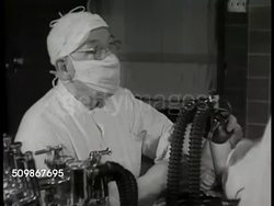 1948: HOSPITAL OPERATION: Wall clock 8:05. Patient into operating room, team of surgeon, technicians, nurses. Surgical light. Female patient w/ oxygen mask being held on face. Male anesthetist nods to surgeon. Operation in progress, obscured Instructional Video