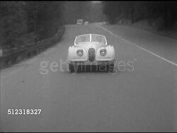 1953: SPORTS CARS: JAGUAR: LA WS (POV Back of moving car) 1953 Jaguar XK-120 convertible driving down road, shaky taking road bumps. MS 'Cass' driving. WS (POV Passenger side) approaching, passing sedan & under old arched overpass. Instructional Video