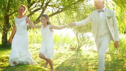 Flower girl holding hands with bride and groom under sunny tree Stock Footage