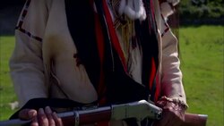 Older male Native American Indian wearing traditional clothing and feathered headdress holding gun with smoke behind Stock Footage