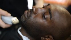 Customer reclines in local barbershop while barber trims his neck and beard with electric clippers Stock Footage