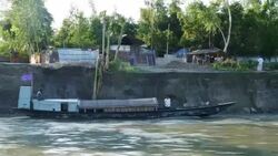 View from a speedboat of a steeply eroded river bank in Bangladesh with children playing and traditional wooden boats ferrying passengers and goods  Stock Footage