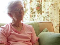 A static shot of an elderly woman in a pink blouse talking on a yellow couch. She has grey hair and circular glasses. The couch has green pillows. Stock Footage