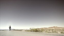 Young skater walks away down desert road carrying skateboard (slow-motion) Stock Footage