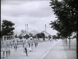 1942: MYSORE, INDIA: VS Male workers dressed in pants, some in shorts, walking down wide street in industrial complex, buildings & tall stacks BG.  No cars, Colonialism, migrants, labor, skilled, unskilled, Sikh, work. Instructional Video