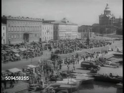 1937: HELSINKI, FINLAND: HA WS Central Railway Station landmark. HA WS Kauppatori (market square) w/ vendors, people shopping, Uspenski Cathedral BG. STREET SCENE: People on sidewalk, man on bicycle FG. People on station sidewalk by passenger train. Instructional Video