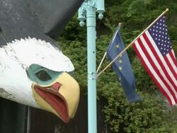 "CU of head of bald eagle wooden statue, Alaskan state flag and national flag of USA flying in the wind behind, on corner of Front Street, Grant Street and Water Street, Ketchikan, Ketchikan Borough, Alaska." Stock Footage