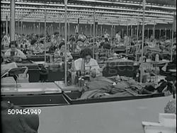 1963: GARMENT FACTORY WORK:  PAN Large room w/ women working at sewing machines. VS Rows of sewing machines w/ women working, bins for completed piece work on other side of table. Apparel Industry, assembly, manufacturer, Made in the USA. Instructional Video