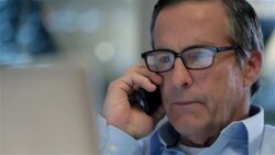 Multitasking businessman scans laptop-computer screen, talks on smart-phone in private corporate office (dolly shot) Stock Footage