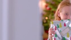 Cute young girl holds present up for camera in front of Christmas tree (dolly-shot) Stock Footage