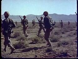 1950s WS Shockwave blowing gust of sand into soldiers / men climbing out of trenches w/ rifles & advancing towards ground zero / PAN up mushroom cloud at Camp Desert Rock at Nevada Test Site / Nye County, Nevada, USA Stock Footage
