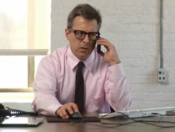 Medium shot. Dolly to the right around man sitting at desk, talking on cell phone, and working at computer Stock Footage
