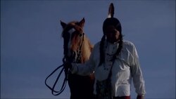 Older male Native American Indian with feather in hair walking brown horse over grassy ridge holding arm up to sky Stock Footage