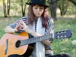 Young woman wearing hat sitting in park outdoors, playing guitar. Stock Footage