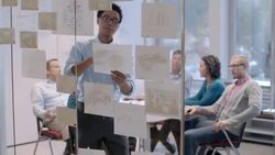Hip young businessman leads meeting, tapes pie-chart to glass-wall of office conference-room (dolly shot) Stock Footage