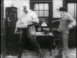 B/W 1916 man (Charlie Chaplin) hiding behind table during pie fight + men throwing pies / 2 shots Stock Footage