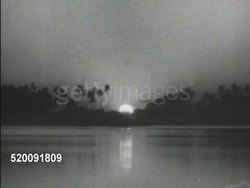 1934: TROPICAL SUNSET: XWS Foliage & tall palm trees in silhouette on opposite water bank, thin smoke rising from darkness, sun setting, moving down behind trees.   River, lake, waterway, island, rainforest. Instructional Video