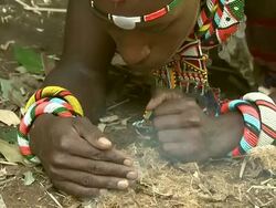 Medium - A Maasai man lies on the ground and blows on a pile of smoldering grass / Kenya Stock Footage
