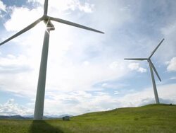 Wind turbines create electrical energy above meadow, mountains Stock Footage