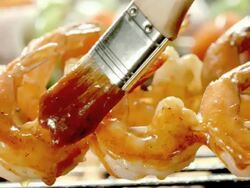 ECU basting brush swipes barbecue sauce over three shrimp on skewers on open flame grill Stock Footage