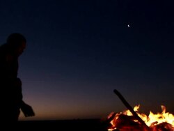 Silhouetted medium shot of person sitting next to camp-fire, standing up, pulling out burning log and throwing it Stock Footage