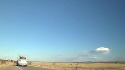 Semi truck carries a backhoe on a trailer and drives down a rural highway and passes a farm tractor discing a field. Stock Footage