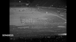 1950: COLLEGE FOOTBALL: NIGHT: Two fingers of male hand holding football vertical, male leg kicking ball. HA Football game in progress, players running up field, kick-off. WS Player running, tackled. Fans in stadium. WS Outdoor flood lighting. (PB) Instructional Video