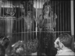 Fairground attraction in France 1947: A man dressed as a colonialist presents a cage containing two Kanak people. One of them performs fire-breathing and the other dances. Stock Footage