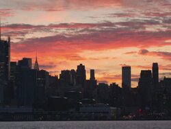 Shot of the Midtown Manhattan Skyline in the late afternoon.  The sky is lit up with oranges, pinks, and purples. The Empire State Building, Chrysler Building, and New York Times Building are visible. A ferry goes along the Hudson River Stock Footage