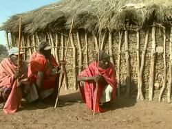 Long Shot - Three Maasai tribesmen socialize as they squat outside a hut / Kenya Stock Footage