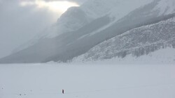 Distant person crosses frozen lake to ice fishing shack, mountains Stock Footage