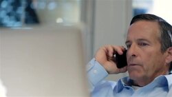 Older businessman on smart-phone puts on glasses and leans forward to inspect info on laptop-computer screen (dolly shot) Stock Footage