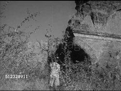 1953: OLD GOA, INDIA: Unidentified boy (8,9 years old) playing harmonica near ruins, standing up, walking away. MS TRACKING Boy walking by tall vegetation, TU WS Crumbling wall sections BG. Instructional Video