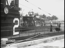 1940: SUBMARINES: MS US Submarine Base sign. XWS Pacific dock & ship w/ palm trees FG. Submarine (7) moving along side, behind docked sub (2), officers watching from deck. Sailors moving torpedo along dock near submarine. Instructional Video